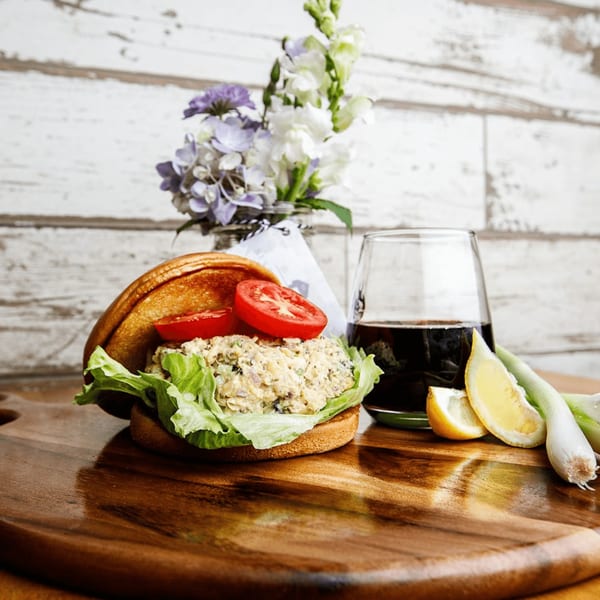 Roast's chicken salad sandwich with lettuce and tomato, a drink, and flowers on a wooden board.