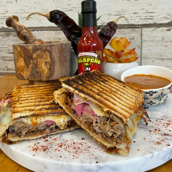 Two halves of a grilled panini sandwich with pulled pork, cheese, and pickled onions, served with a side of soup.