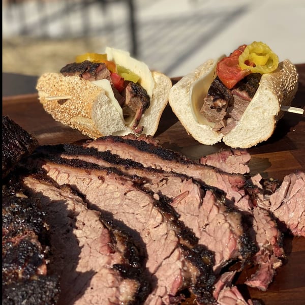 Sliced Roast brisket with two brisket sandwiches topped with cheese, peppers, and tomatoes.