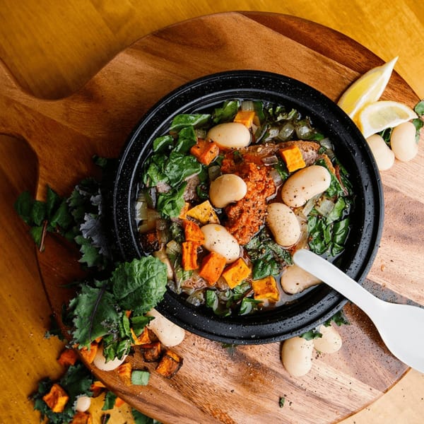 Hearty stew with white beans, sweet potatoes, greens, and a dollop of red sauce, on a wooden board.