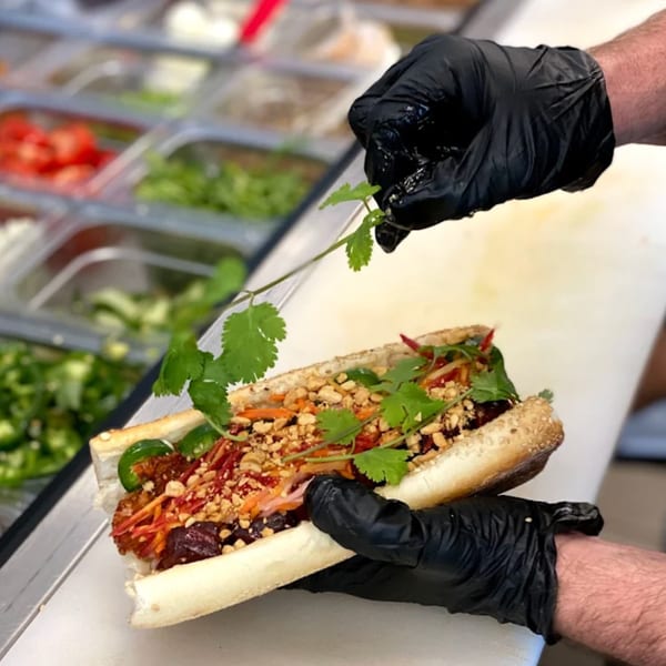 Gloved hands garnishing a Roast sandwich with cilantro, peanuts, and vegetables on a counter.