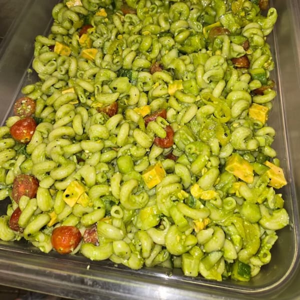 Close-up of green pasta salad with cherry tomatoes and cheese cubes in a metal serving tray.