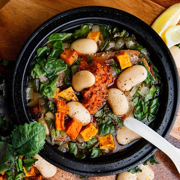 Hearty American stew with white beans, sweet potatoes, and greens in a black bowl with lemon wedges.