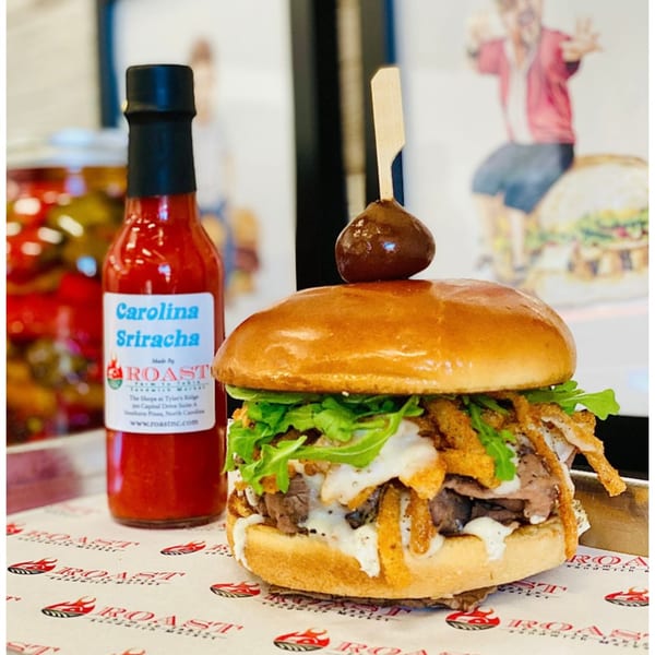Roast's Carolina Sriracha bottle next to a roast beef sandwich with arugula, crispy onions, and sauce.