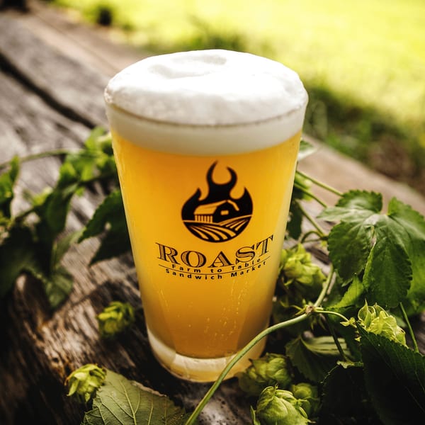 A glass of Roast beer with a foamy head, surrounded by green hops on a wooden surface.